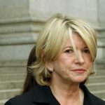 A woman with blonde hair in a black outfit, standing outdoors with a serious expression