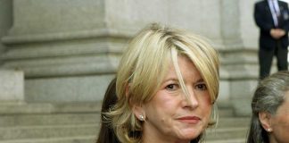 A woman with blonde hair in a black outfit, standing outdoors with a serious expression